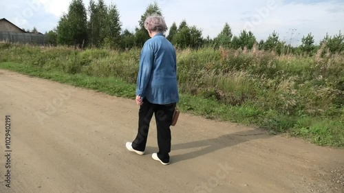 An elderly woman walks with a string bag along a flat village road in the summer