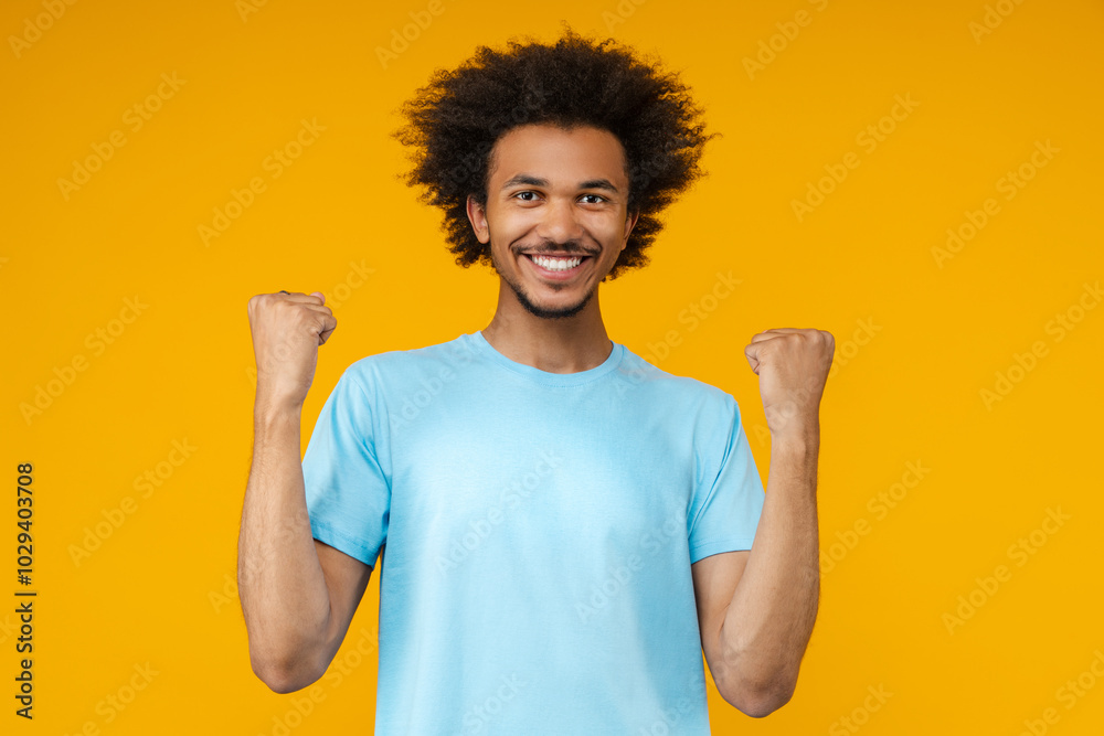 Portrait of happy smiling handsome young African American man in basic light blue t-shirt posing in studio making winners gesture clenching his fists, isolated over bright colored yellow background Portrait of happy smiling handsome young African American man in basic light blue t-shirt posing in studio making winners gesture clenching his fists, isolated over bright colored yellow background