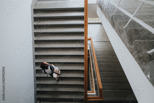 A student with a backpack ascends a wide staircase in a well-lit, modern building, symbolizing education and progress. The architectural lines create a striking visual perspective. photo
