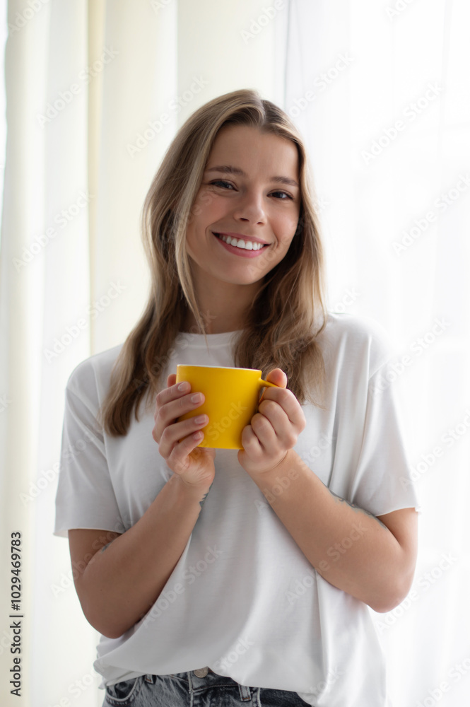 Lovely young woman stands by a bright window, smiling while holding a yellow mug. She wears a white t-shirt, enjoying a peaceful moment in a minimalist, light-filled room, evoking warmth and calm. Lovely young woman stands by a bright window, smiling while holding a yellow mug. She wears a white t-shirt, enjoying a peaceful moment in a minimalist, light-filled room, evoking warmth and calm.