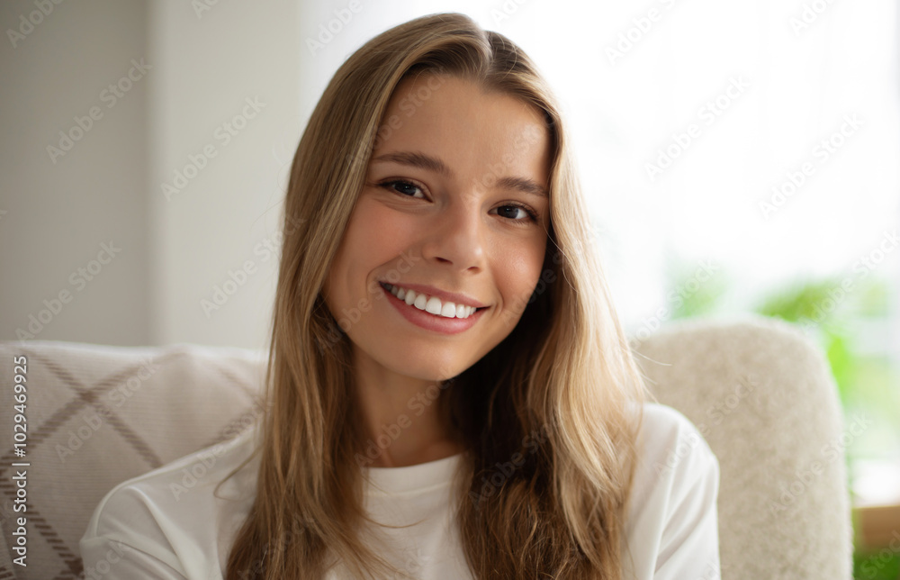 Portrait of attractive smiling young woman with long blonde hair sitting indoors, wearing casual white shirt. The background is bright and cozy, featuring white chair and soft natural lighting Portrait of attractive smiling young woman with long blonde hair sitting indoors, wearing casual white shirt. The background is bright and cozy, featuring white chair and soft natural lighting
