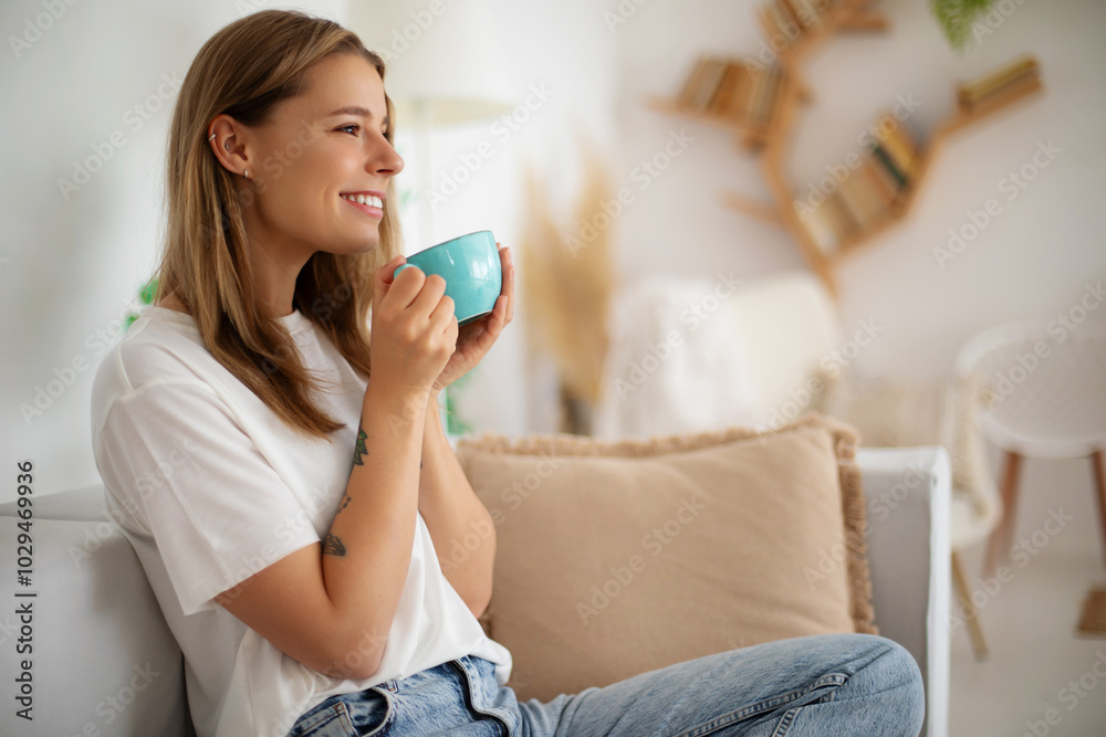 Happy young woman relaxes and sits comfortably on a sofa, smiling while holding a blue cup. She wears a casual white t-shirt and jeans in a cozy, modern living room with soft pillows, warm lighting Happy young woman relaxes and sits comfortably on a sofa, smiling while holding a blue cup. She wears a casual white t-shirt and jeans in a cozy, modern living room with soft pillows, warm lighting