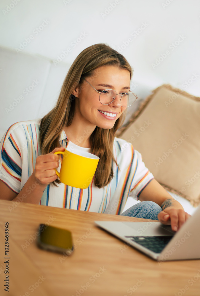 Lovely young woman sits at a wooden table with a laptop, smiling and holding a yellow mug. She wears a casual striped shirt and glasses, working in a cozy, bright home office setting with plants.
