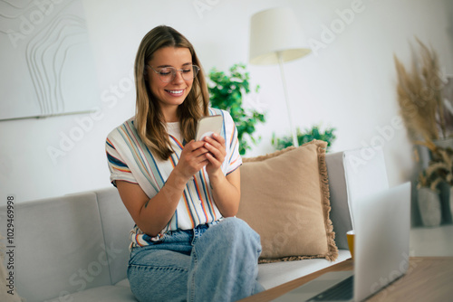 Young woman sits comfortably on a sofa, casually dressed in jeans and a striped shirt. She smiles while using her smartphone, with a laptop on the table in front of her in cozy, modern living space