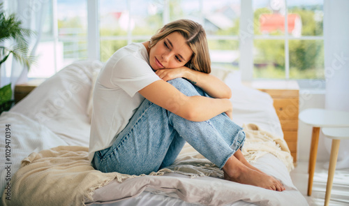Obraz na plátně Depressed woman sits on her bed with her knees hugged to her chest, appearing sad or reflective
