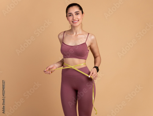 Weight Loss And Healthy Lifestyle Concept. Smiling slim woman measuring her waist with yellow tape over pastel studio background. Fit young lady in perfect shape, happy with her body