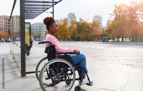 Lack of public transport for disabled people. Young black paraplegic woman in wheelchair feeling upset, waiting on bus stop in autumn, cannot board vehicle suitable for handicapped persons, copy space