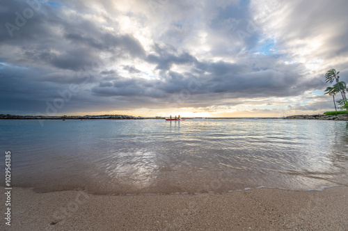 As the sun sets, two individuals paddle in a kayak on a peaceful beach while dark clouds loom overhead, reflecting the day's last light on the water's surface.