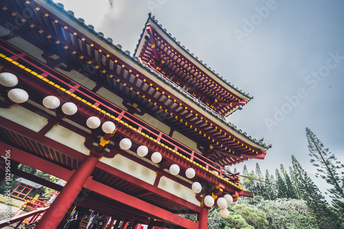The structure features intricate designs with vibrant red and gold accents, surrounded by lush greenery and soft clouds, creating a tranquil atmosphere.