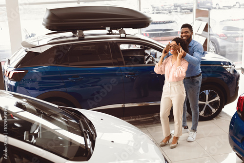 Own Car. African Amrerican Husband Covering Wife's Eyes Showing Her New Auto In Dealership Store. Empty Space photo