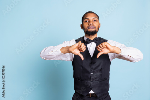 Discontent unhappy bellhop gives a thumbs down symbol on camera, showing rejection and disagreement. Dissatisfied hotel staff with suit and tie does a dislike sign, feeling displeased. photo