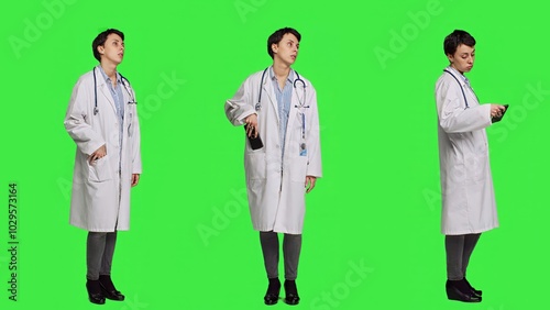 Woman specialist looking at phone watch and waiting for patients to arrive at exam appointments, greenscreen backdrop in studio. General practitioner in white coat acting impatient. Camera A.