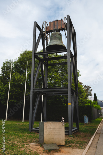 Large bronze bell housed in a metal tower surrounded by greenery on a cloudy day in an open area photo