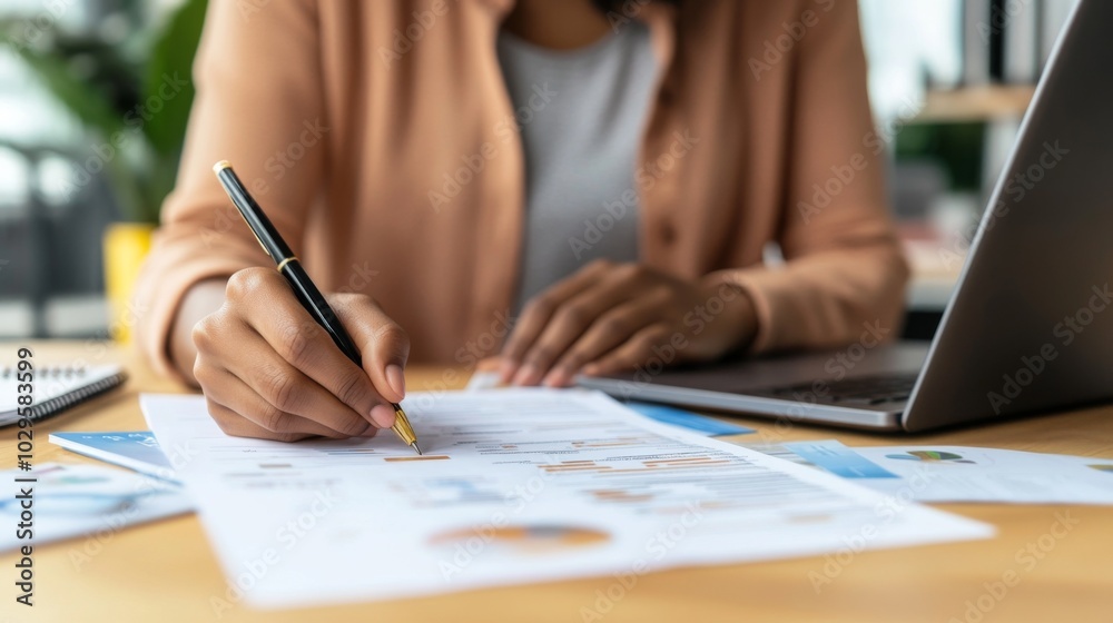Person is analyzing business charts and graphs on paper, using a pen, with a laptop nearby, emphasizing productivity and data organization.