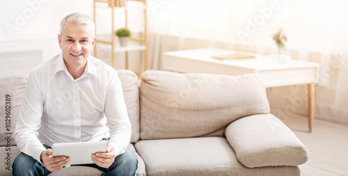 Portrait of senior sman sitting on a modern couch, holding digital tablet and smiling broadly at the camera photo