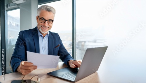 Happy busy older professional businessman entrepreneur using laptop holding bill corporate document in office. 50 years old man executive manager or business owner reading paper working on compute