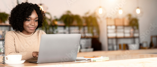 Young positive black lady drinking tea and using laptop, cafe interior, empty space