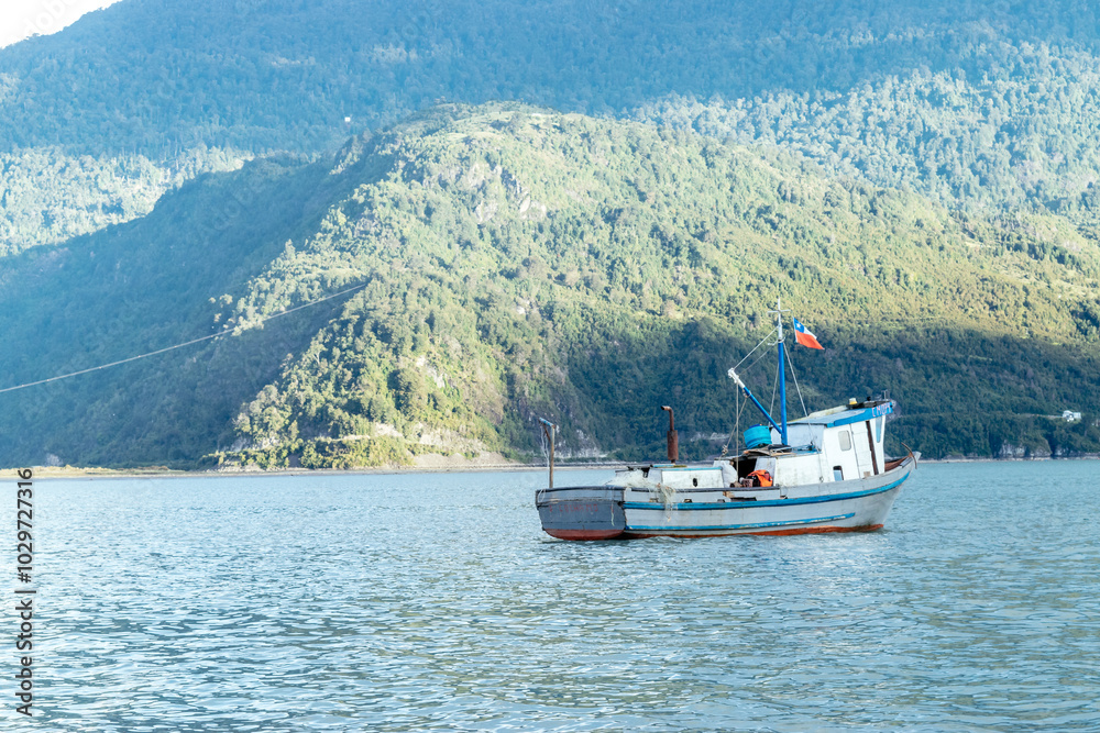 Small fishing boat with chilean flag is floating on calm water near a mountain covered by green
