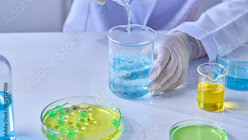 Photo was shot from close high angle view at white laboratory table, which displaying various lab glassware contain colorful liquid and gel ball, a scientist pours blue essence into glass beaker