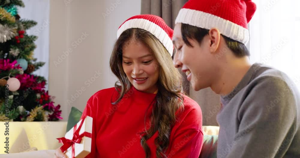 Asian couple wearing Santa hats, smiling while the woman excitedly unwraps a Christmas gift. Both enjoying the joyful moment together in a cozy living room, surrounded by festive holiday decorations. Asian couple wearing Santa hats, smiling while the woman excitedly unwraps a Christmas gift. Both enjoying the joyful moment together in a cozy living room, surrounded by festive holiday decorations.