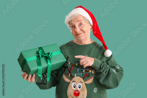 Senior woman in Santa hat pointing at Christmas gift on green background