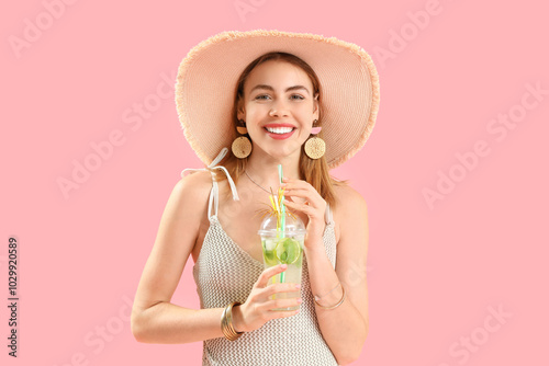 Beautiful young woman in wicker hat with glass of tasty mojito on pink background