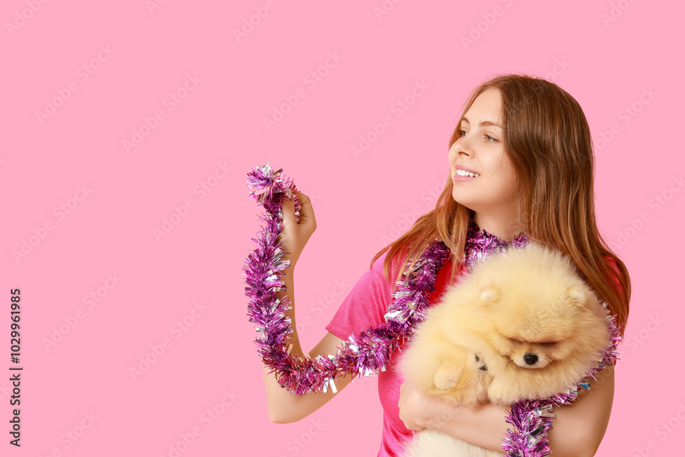 Young woman with cute Pomeranian dog and Christmas tinsel on pink background