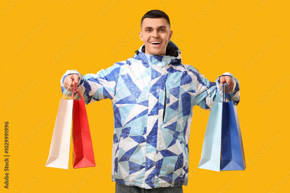 Young man in stylish puffer jacket with shopping bags on yellow background Young man in stylish puffer jacket with shopping bags on yellow background