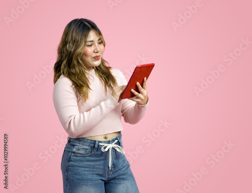 Young asian woman in casual clothing, holding and looking at a tablet computer. Portrait on a simple pink background with studio light. Side view