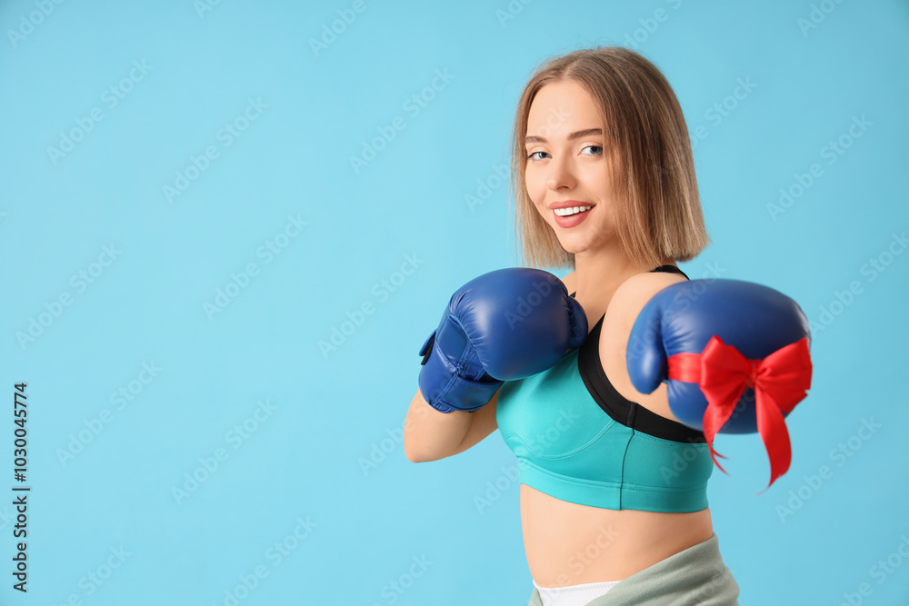 Sporty young woman in boxing gloves with Christmas ribbon on blue background Sporty young woman in boxing gloves with Christmas ribbon on blue background