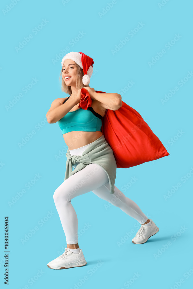 Obraz premium Sporty young woman in Christmas hat carrying Santa Claus bag on blue background