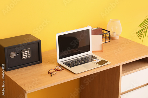 Modern laptop and safe on table near yellow wall