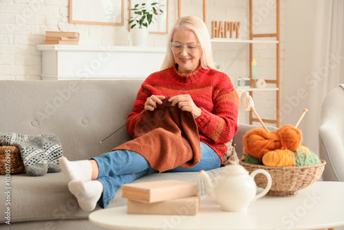 Mature woman knitting sweater with needles on sofa at home