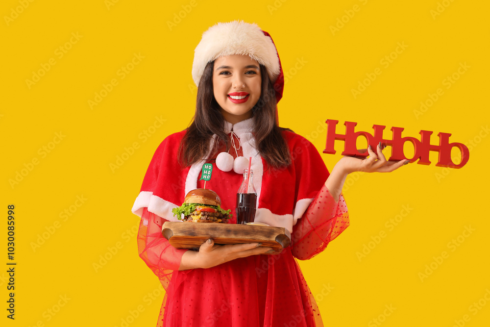 Pretty young woman in Santa costume with tasty burger, cola and HOHOHO word on yellow background Pretty young woman in Santa costume with tasty burger, cola and HOHOHO word on yellow background