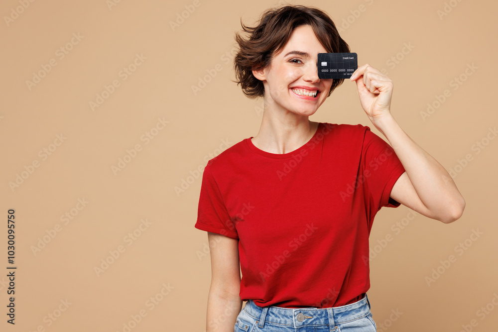 Young smiling happy woman she wears red t-shirt casual clothes hold in hand cover eye face with mock up of credit bank card isolated on plain pastel light beige background studio. Lifestyle concept. Young smiling happy woman she wears red t-shirt casual clothes hold in hand cover eye face with mock up of credit bank card isolated on plain pastel light beige background studio. Lifestyle concept.