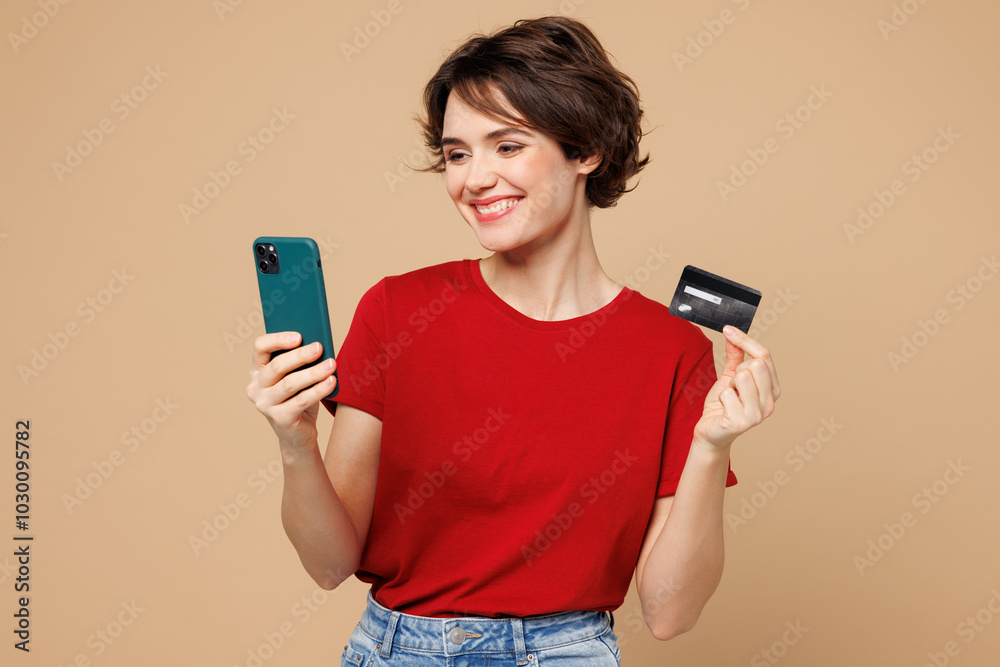 Young woman wear red t-shirt casual clothes using mobile cell phone hold credit bank card doing online shopping order delivery booking tour isolated on plain beige background studio Lifestyle concept Young woman wear red t-shirt casual clothes using mobile cell phone hold credit bank card doing online shopping order delivery booking tour isolated on plain beige background studio Lifestyle concept