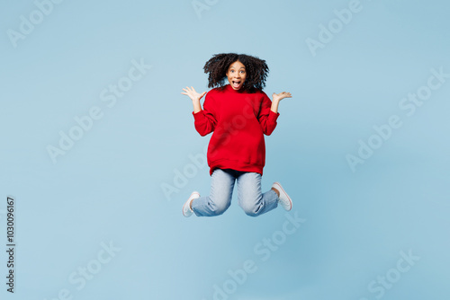 Full body shocked surprised little kid teen girl of African American ethnicity wear red sweatshirt jump high spread hands isolated on pastel plain light blue background. Childhood lifestyle concept.