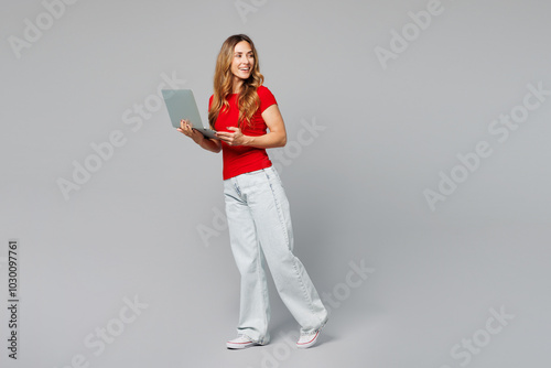 Full body young smiling happy IT woman she wear red t-shirt casual clothes hold use work on laptop pc computer look aside on area isolated on plain grey background studio portrait. Lifestyle concept. photo