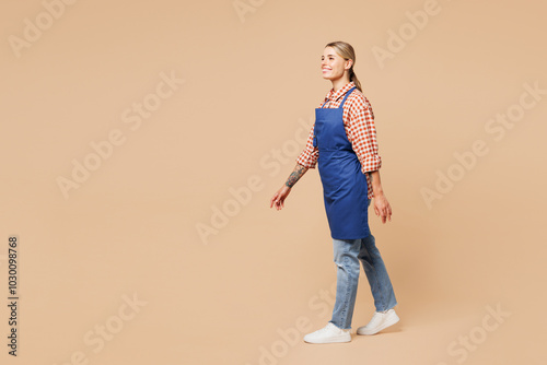 Full body side profile view young woman barista bartender barman employee she wear blue apron red shirt work in coffee shop walk go isolated on plain beige background. Small business startup concept. photo