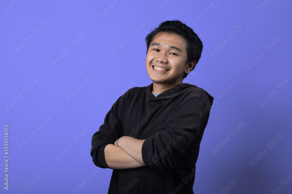 An Asian man is smiling with his arms crossed, standing against a purple background. He is wearing a dark hoodie and exudes confidence and warmth in a casual, relaxed pose An Asian man is smiling with his arms crossed, standing against a purple background. He is wearing a dark hoodie and exudes confidence and warmth in a casual, relaxed pose