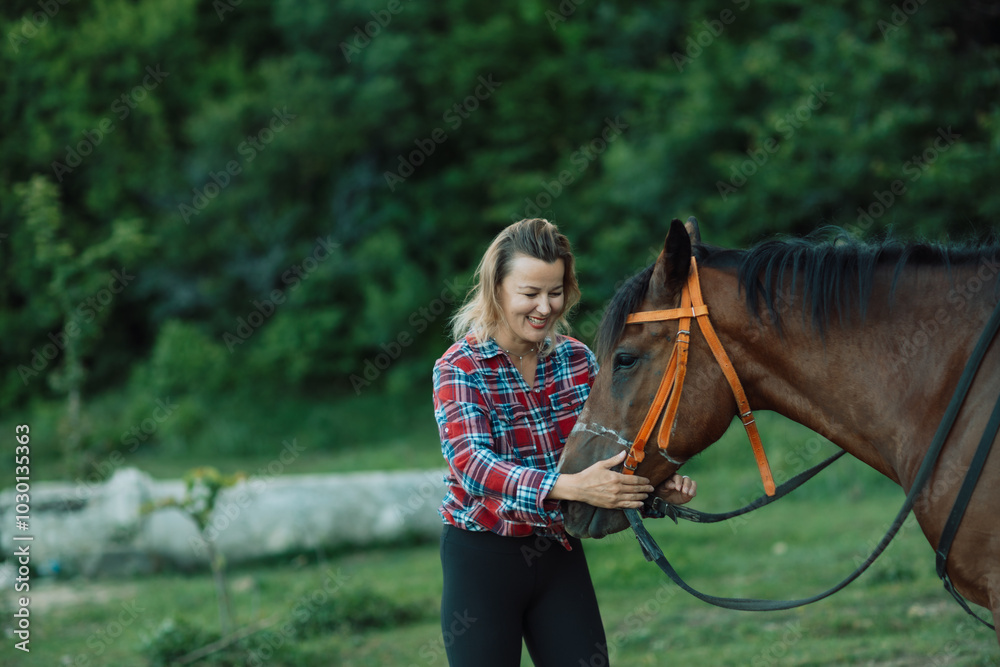 Obraz premium Happy blonde with horse in forest. Woman and a horse walking through the field during the day. Dressed in a plaid shirt and black leggings.