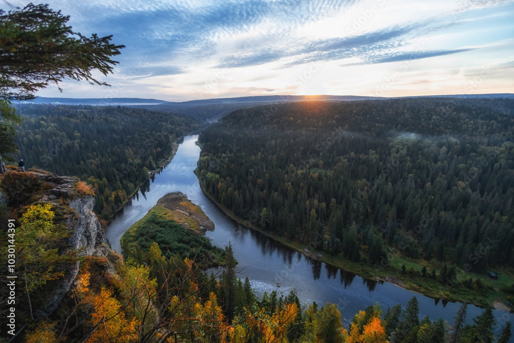 Obraz premium The sun sets behind rolling hills, casting a warm glow over a winding river embraced by vibrant autumn trees, creating a peaceful avenue of natureвЂ™s beauty