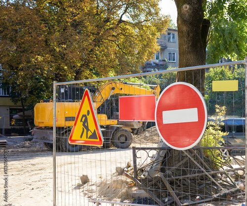 Construction equipment on the site, a fence with a stop sign, work is underway. A tractor levels the ground and carries sand on a rack, builders build a building, parking on the street