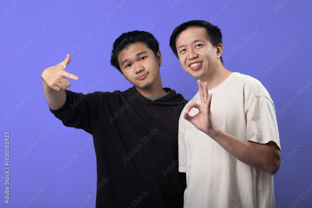 Two Asian men are posing confidently against a purple background. The man on the left is pointing at the camera, while the man on the right is making an Two Asian men are posing confidently against a purple background. The man on the left is pointing at the camera, while the man on the right is making an