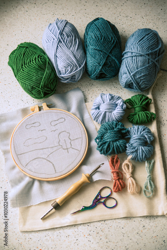 Sketch of a landscape in round embroidery frame with a bunch of woolen skeins and some instruments for tufting.