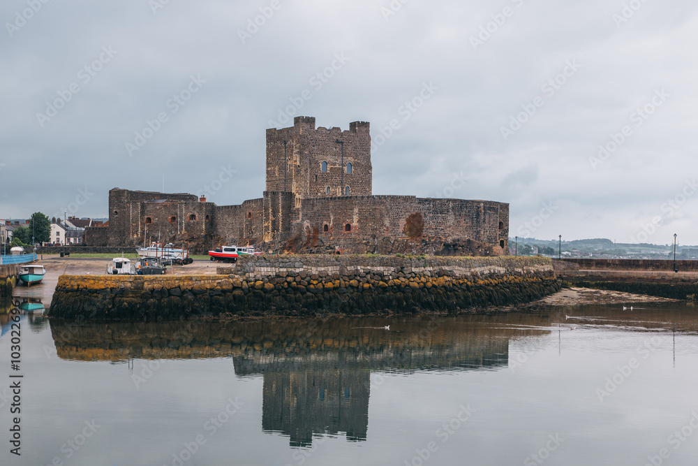 Fototapeta premium Carrickfergus Castle reflected in calm waters, with boats docked along the stone walls. The medieval fortress stands against a cloudy sky in Northern Ireland harbor