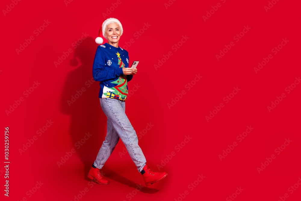 Obraz premium Full body portrait of pretty aged lady hold smart phone walk empty space new year xmas isolated on vibrant red color background