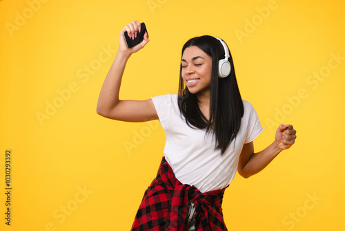 Positive young african american girl woman female student listening enjoyed music song radio dancing having fun holding smart mobile phone isolated on yellow background. Technology concept