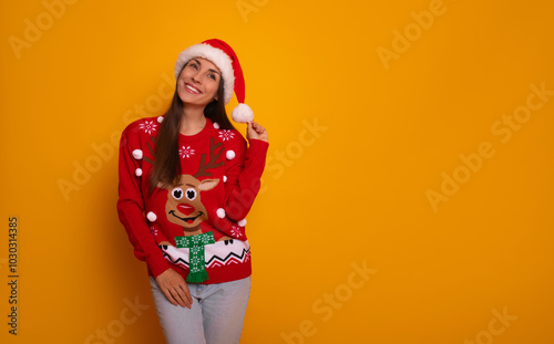 Cheerful woman in a festive Santa hat and a red Christmas sweater with a reindeer design smiles joyfully against a vibrant orange background. Holiday-themed promotions and cheerful winter campaigns photo