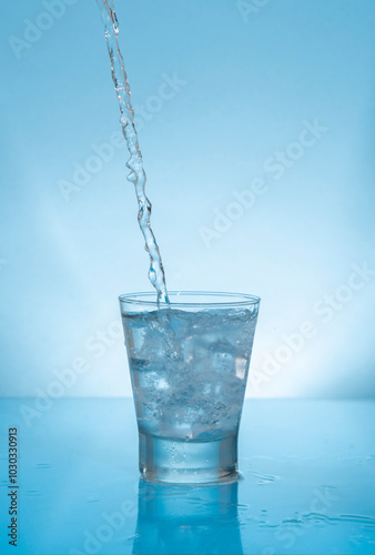 Water stream splashing on glass cup filled with water and ice on wet reflective blue surface
